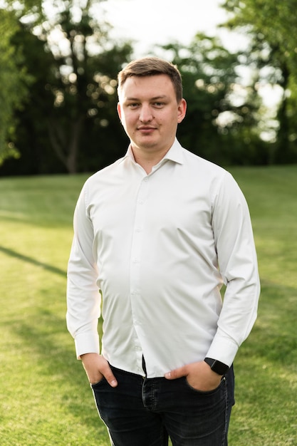 Portrait d'un jeune homme en chemise blanche marchant dans le parc au coucher du soleil. Un Européen regarde la caméra. homme sur le fond de la pelouse du parc.