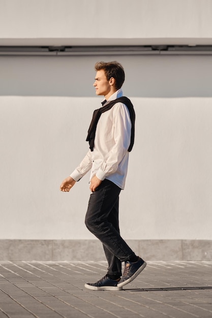 Portrait d'un jeune homme en chemise blanche et pull drapé sur les épaules marche dans la rue