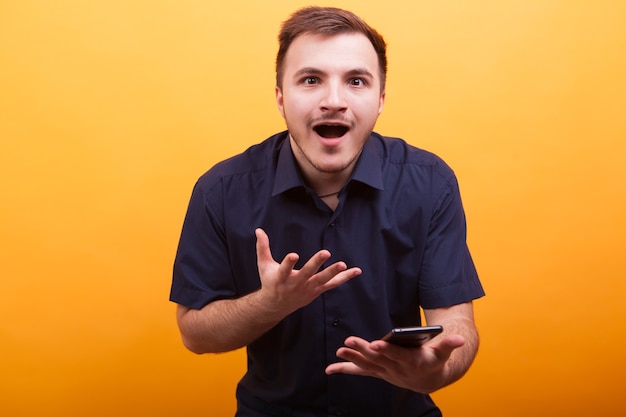 Portrait de jeune homme choqué en chemise bleue tenant un téléphone portable isolé sur fond jaune. Visage surpris