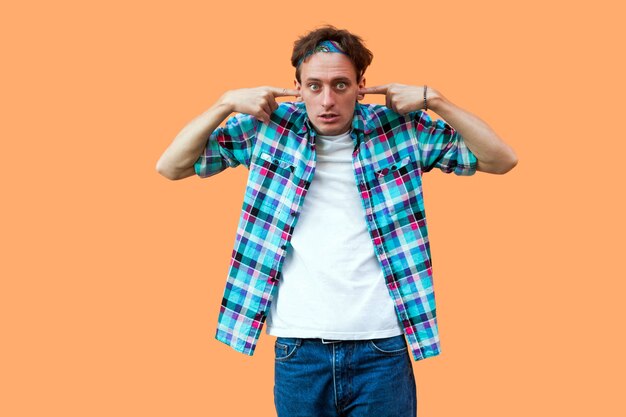 Portrait D'un Jeune Homme Choqué Ou Confus En Chemise à Carreaux Bleu Décontractée Et Bandeau Debout, Mettant Le Doigt Sur Ses Oreilles Et Regardant La Caméra. Tourné En Studio Intérieur, Isolé Sur Fond Orange.