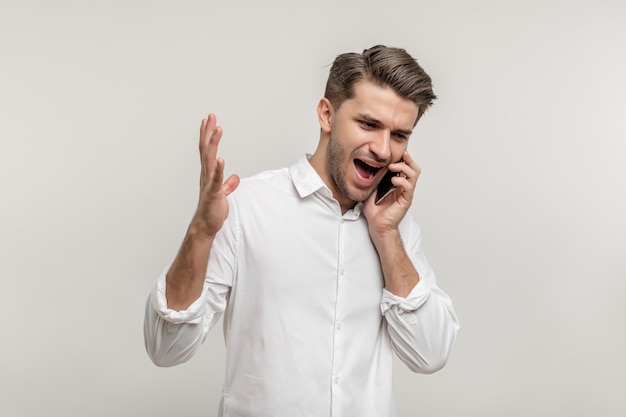 Portrait d'un jeune homme en colère en chemise blanche, parler au téléphone mobile isolé sur fond blanc