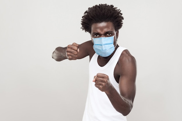 Portrait d'un jeune homme en colère portant une chemise blanche avec un masque médical chirurgical debout avec des poings de boxe et prêt à attaquer ou à se défendre. tourné en studio intérieur isolé sur fond gris.