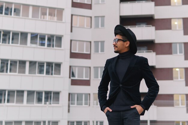 Portrait d'un jeune homme cool en costume noir et chapeau marchant sur le bâtiment d'arrière-plan