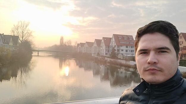 Portrait d'un jeune homme debout au bord d'une rivière dans la ville contre le ciel au coucher du soleil