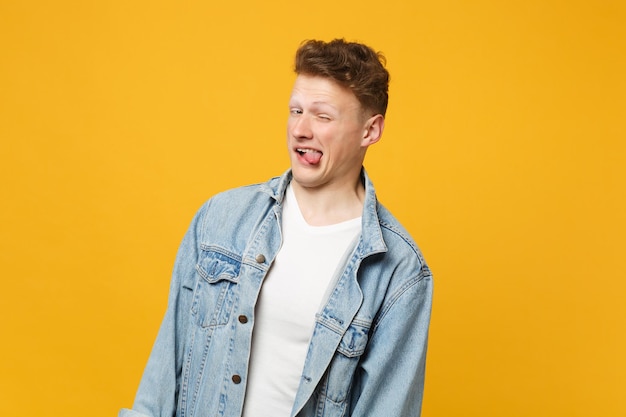 Portrait d'un jeune homme drôle dans des vêtements décontractés en denim montrant la langue et clignotant isolé sur fond de mur orange jaune en studio. Émotions sincères des gens, concept de style de vie. Maquette de l'espace de copie.