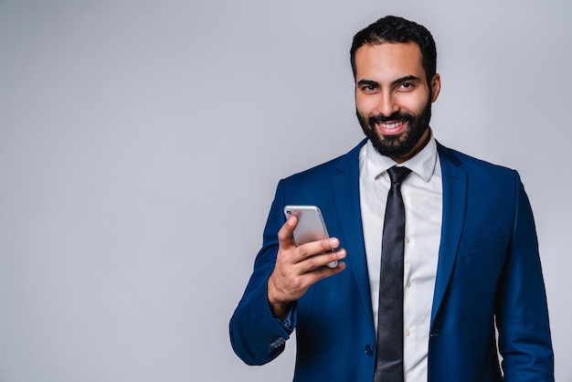 Portrait d'un jeune homme du Moyen-Orient souriant en costume d'affaires utilisant un téléphone portable isolé sur un fond gris