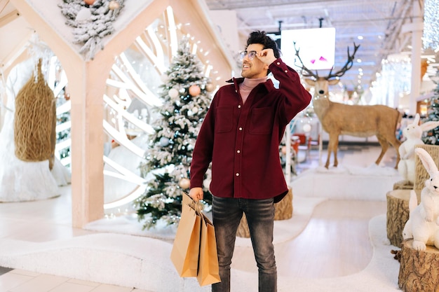 Portrait d'un jeune homme élégant tenant des sacs en papier et souriant regardant loin tenant un temple de spectacles debout dans le hall de célébrer le centre commercial