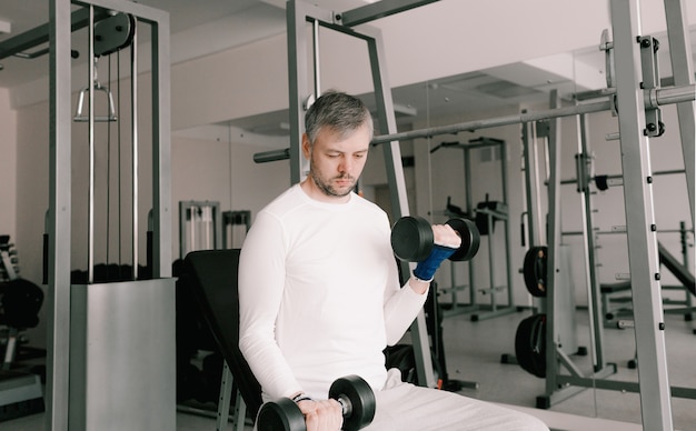 Portrait d'un jeune homme faisant des exercices physiques, entraînement des biceps avec des haltères dans la salle de gym, une copie de l'espace. Dans un T-shirt blanc.