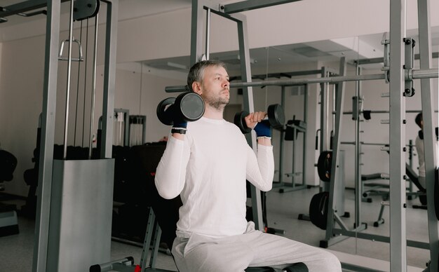 Portrait d'un jeune homme faisant des exercices physiques, s'entraînant avec des haltères assis sur un banc dans la salle de gym. Dans un T-shirt blanc. mode de vie sain