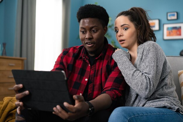 Portrait d'un jeune homme et d'une femme américains assis sur un canapé gris à la maison regardant quelque chose u