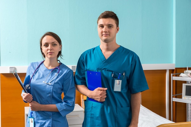 Portrait de jeune homme et femme médecin en uniforme avec phonendoscope sur son cou tenant des presse-papiers en se tenant debout dans la salle d'hôpital. Concept de soins de santé