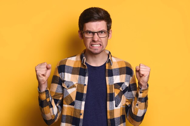 Portrait d'un jeune homme furieux en colère dans des lunettes devenant fou de stress, de surmenage et de problèmes, mains dans les poings, geste de colère, isolé sur fond jaune, semble agressif. Notion d'émotions humaines