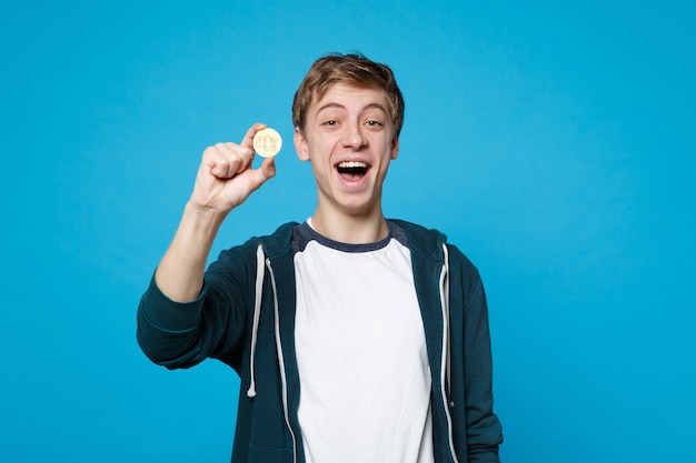 Portrait d'un jeune homme gai dans des vêtements décontractés tenant du bitcoin, future monnaie isolée sur un mur bleu. Les gens émotions sincères, concept de style de vie.