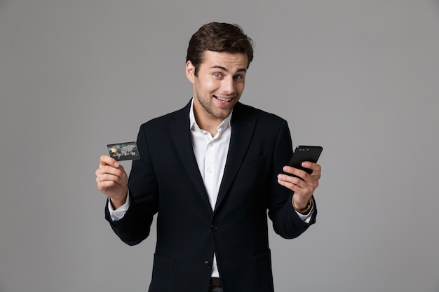 Portrait d'un jeune homme gai habillé en costume isolé sur mur gris, à l'aide de téléphone mobile, montrant la carte de crédit