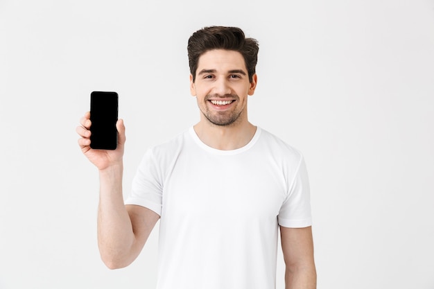 Portrait d'un jeune homme gai portant des vêtements décontractés isolé sur blanc, montrant un téléphone portable à écran blanc