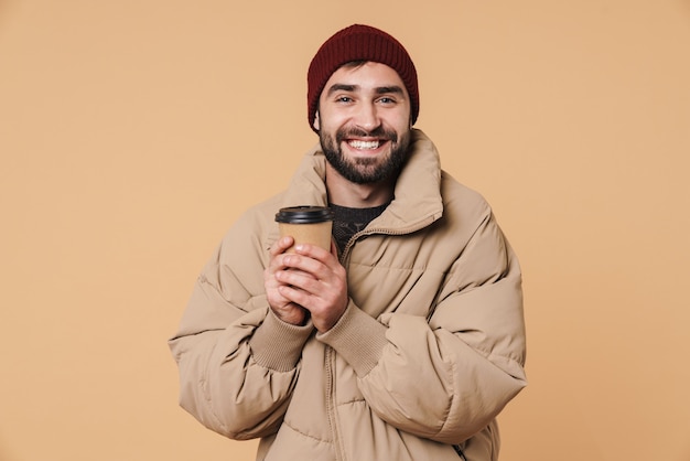 Portrait de jeune homme gai en veste d'hiver et chapeau souriant en buvant du café isolé sur beige