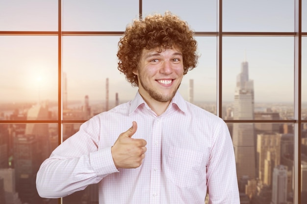 Photo portrait d'un jeune homme heureux aux cheveux bouclés montre le pouce vers le haut fenêtre à carreaux avec vue sur le paysage urbain