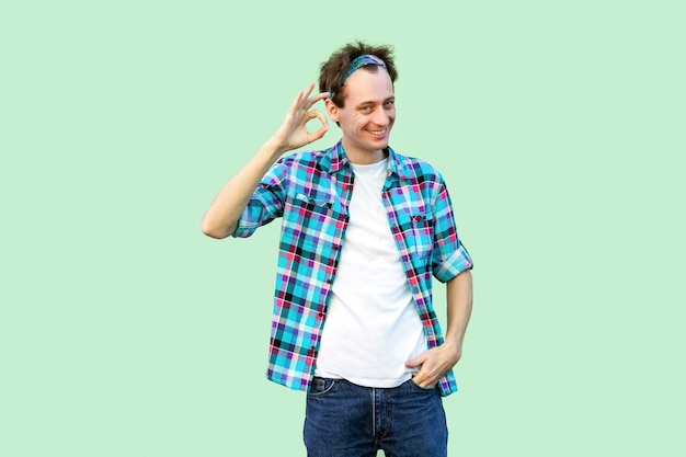 Portrait d'un jeune homme heureux en chemise à carreaux bleu décontractée et bandeau debout avec signe Ok et regardant la caméra avec un sourire à pleines dents. tourné en studio intérieur, isolé sur fond vert clair.