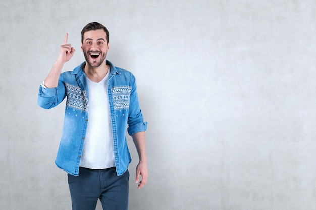 Portrait d'un jeune homme heureux en chemise jeans et pantalon debout avec la bouche ouverte et le doigt pointant vers le haut près du mur de béton. Concept d'avoir une idée. Maquette