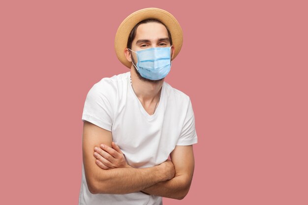 Portrait d'un jeune homme heureux avec un masque médical chirurgical en chemise blanche avec chapeau debout, bras croisés et regardant la caméra avec le sourire. tourné en studio intérieur, isolé sur fond rose.