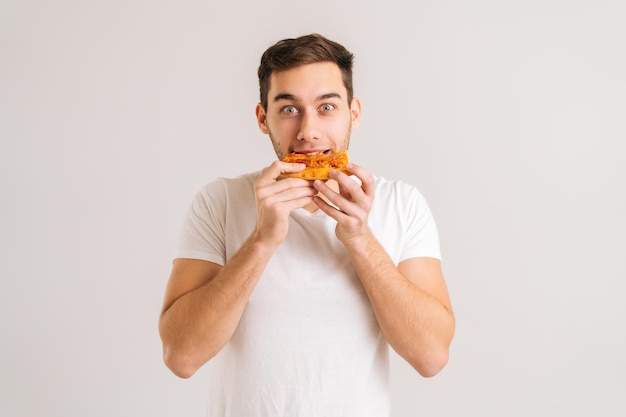 Portrait d'un jeune homme heureux mordant une délicieuse tranche de pizza en regardant la caméra sur fond isolé blanc