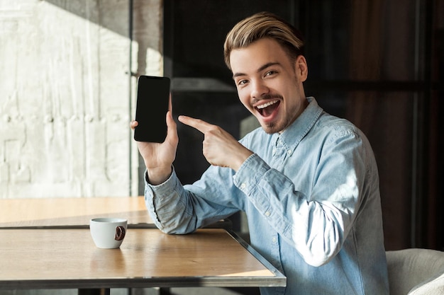 Portrait d'un jeune homme heureux et positif en chemise bleu denim assis dans un café, tenant un téléphone et pointant du doigt avec un sourire à pleines dents et un visage incroyable sur l'écran mobile, bouche ouverte. Intérieur, style de vie