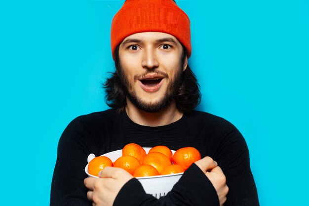 Portrait de jeune homme heureux tenant un bol avec des mandarines sur fond bleu. Portant un chapeau orange et un pull noir.