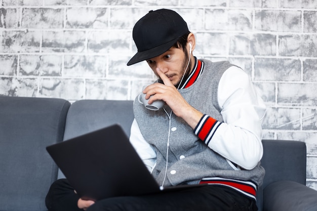 Portrait de jeune homme indépendant, assis sur un canapé avec une tasse de café à la main et un ordinateur portable sur les jambes. Mur de briques grises.