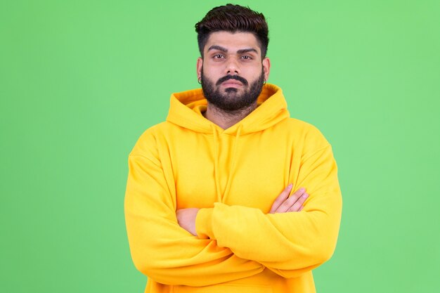 Portrait de jeune homme indien barbu en surpoids portant un sweat à capuche contre chroma key avec mur vert