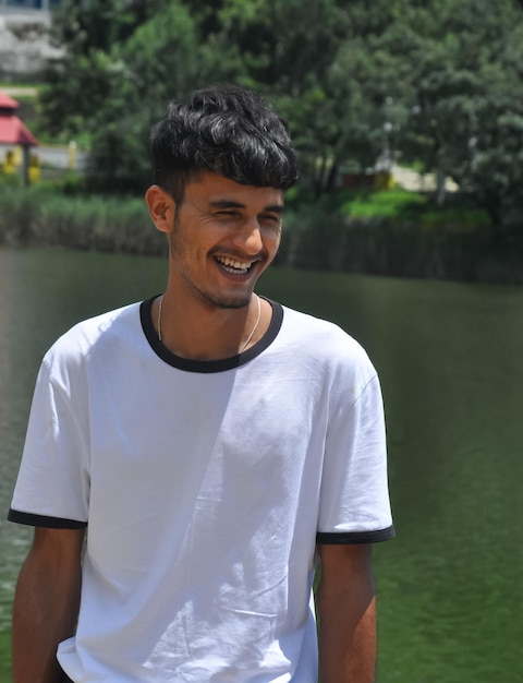 Portrait d'un jeune homme indien debout et souriant à côté du lac avec un t-shirt blanc.