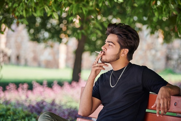 Portrait de jeune homme indien fumeur en t-shirt noir et chaîne de cou en argent assis sur un banc de parc