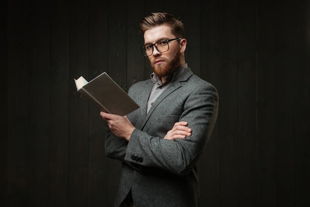 Portrait d'un jeune homme intelligent en costume décontracté debout et tenant un livre isolé sur le fond en bois noir