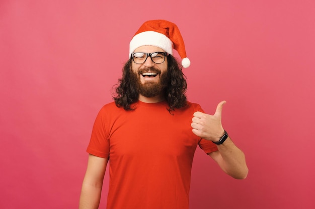 Portrait d'un jeune homme joyeux avec une barbe portant un chapeau de père noël montrant le pouce vers le haut sur fond rose