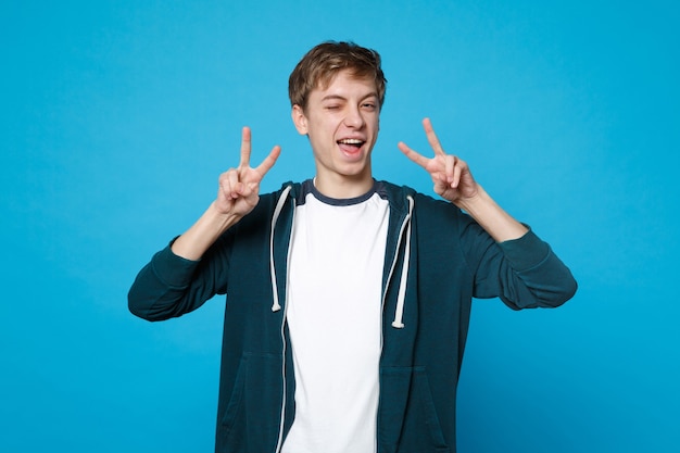 Portrait d'un jeune homme joyeux et joyeux dans des vêtements décontractés clignotant et montrant le signe de la victoire isolé sur le mur bleu.