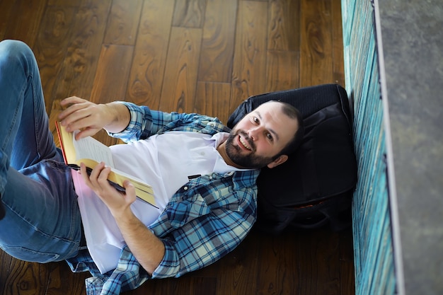 Portrait d'un jeune homme joyeux lisant un livre assis sur le sol dans son salon. Étudiant tenant et lisant un livre.