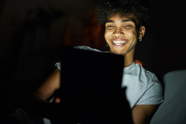 Portrait d'un jeune homme joyeux souriant tout en utilisant un tablet pc la nuit assis dans une pièce sombre à la maison