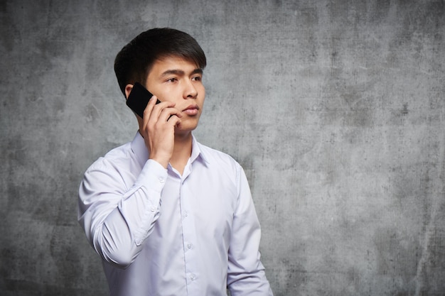 Portrait d'un jeune homme kazakh parlant au téléphone avec une expression faciale sérieuse sur fond gris studio