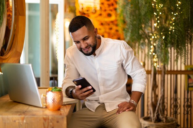 Portrait de jeune homme latin souriant à l'aide de son téléphone portable à l'intérieur d'un bar à cocktails moderne.