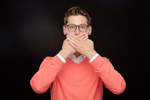 Portrait de jeune homme à lunettes couvrant la bouche avec les mains tout en ne faisant aucun signe parlant, fond isolé