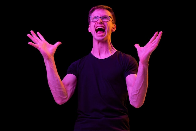 Portrait de jeune homme à lunettes posant avec émotion criant isolé sur fond noir en néon