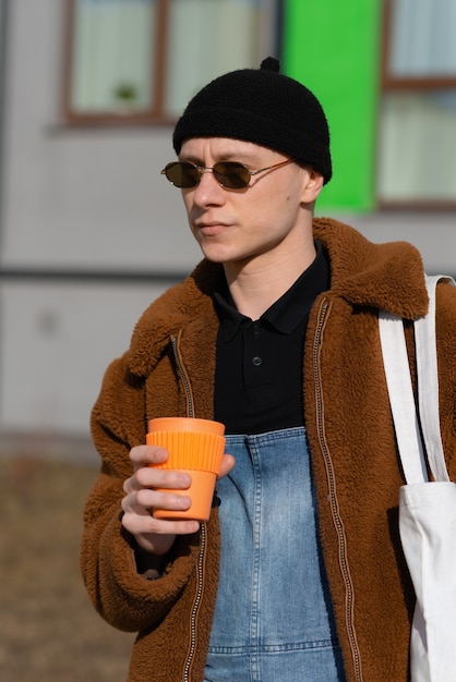 Portrait de jeune homme avec lunettes de soleil et bonnet et tasse de café orange