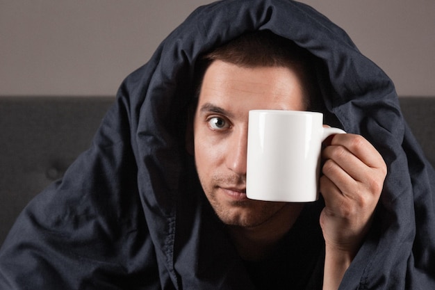 Portrait d'un jeune homme malade buvant du thé au lit malade caucasien assis avec une tasse de boisson chaude dans ses mains avec de la fièvre ne se sentant pas bien à la maison gros plan