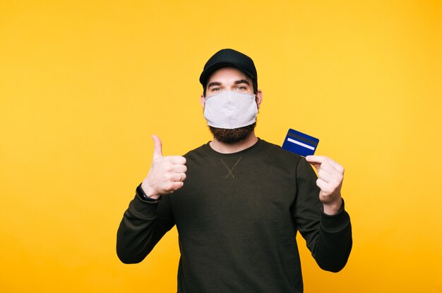 Portrait de jeune homme avec masque facial montrant la carte de crédit et montrant les pouces vers le haut