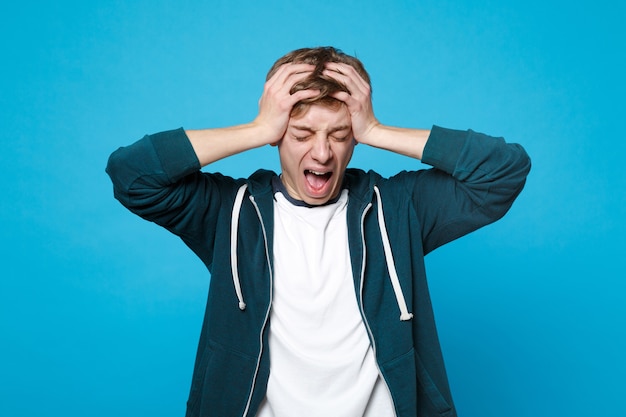 Portrait d'un jeune homme mécontent dans des vêtements décontractés criant, pleurant en mettant les mains sur la tête isolée sur le mur bleu. Les gens émotions sincères, concept de style de vie.