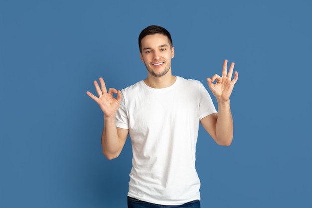 Portrait de jeune homme montrant le signe ok isolé sur le mur bleu du studio