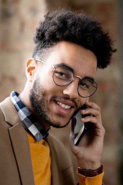 Portrait d'un jeune homme noir gai dans des lunettes et un pull orange utilisant un téléphone portable pour la communication