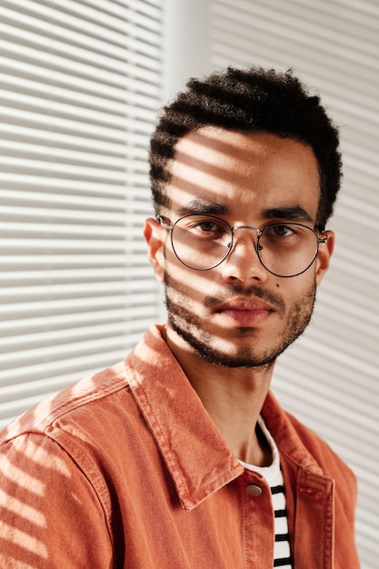 Portrait de jeune homme noir sérieux en lunettes rondes et chemise corail assis contre fermé