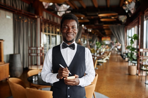 Portrait d'un jeune homme noir en tant que serveur dans un restaurant de luxe souriant à la caméra et tenant un bloc-notes prêt à prendre des commandes.