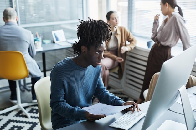 Portrait d'un jeune homme noir utilisant un ordinateur portable sur le lieu de travail avec des personnes discutant en arrière-plan en entreprise