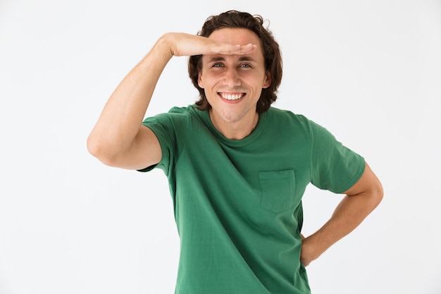 Portrait d'un jeune homme optimiste portant un t-shirt basique souriant et regardant avec sa main sur le front isolé sur un mur blanc